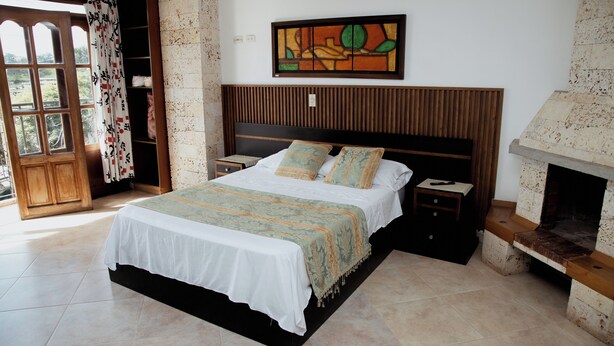 A bedroom with a bed, a nightstand, a wooden headboard, a fireplace, and a window with curtains. - Hotel Portal Guatapé  (Guatapé)