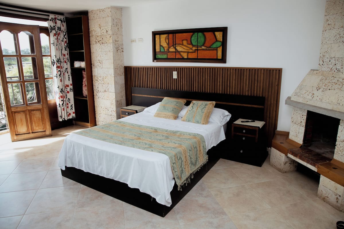 Quarto com cama, mesa de cabeceira, cabeceira de madeira, lareira e janela com cortinas.