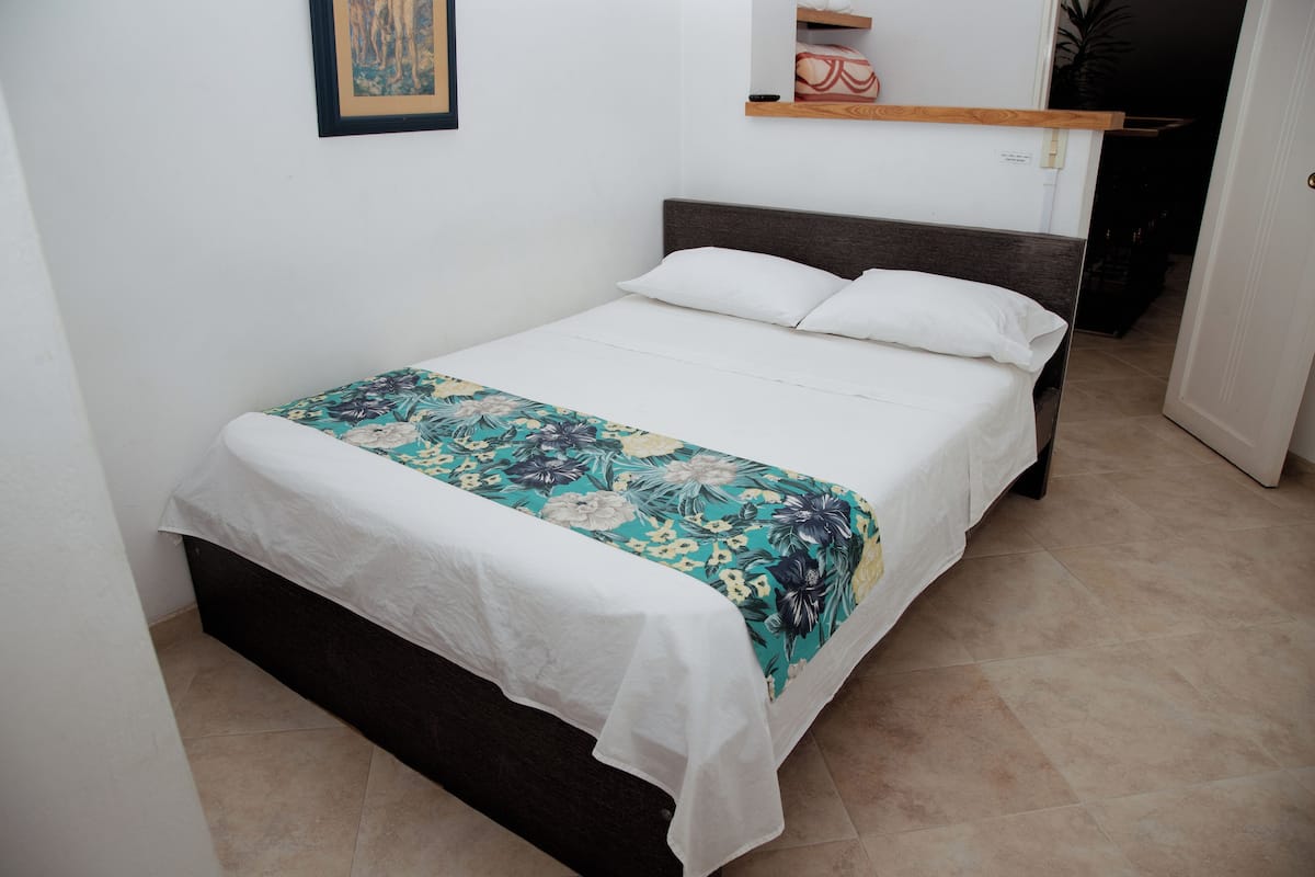 Uma cama bem arrumada com colcha florida e almofadas brancas, um painel de madeira e uma pequena prateleira acima.