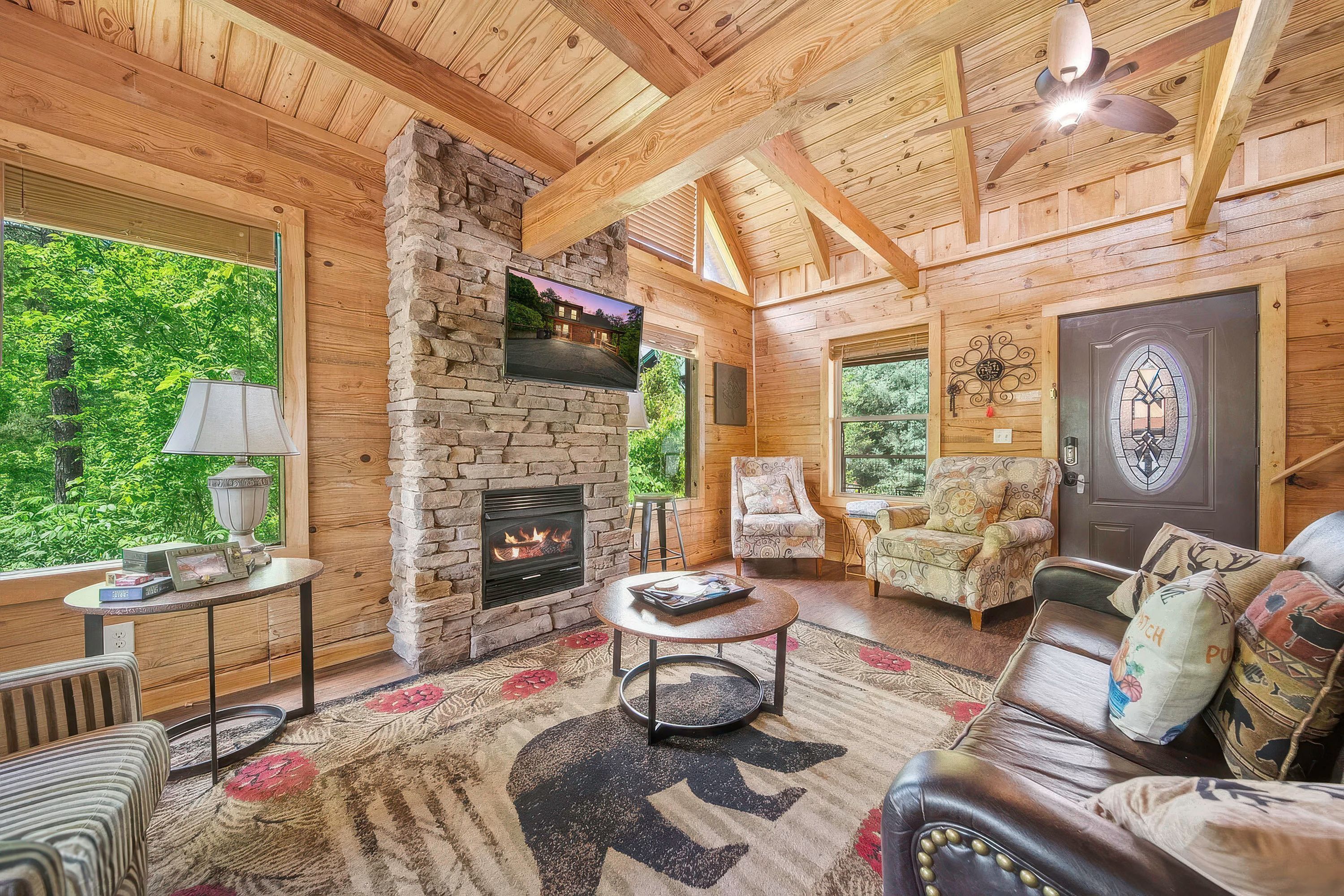 A cozy cabin interior with a stone fireplace, wooden walls, a large window, and a comfortable seating area.