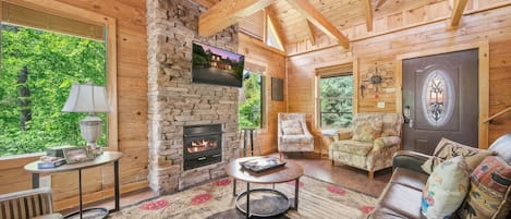 A cozy cabin interior with a stone fireplace, wooden walls, a large window, and a comfortable seating area.