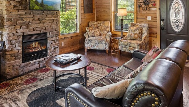 L’intérieur chaleureux d’un chalet avec une cheminée en pierre, des poutres en bois au plafond, un canapé en cuir et deux fauteuils à motifs.