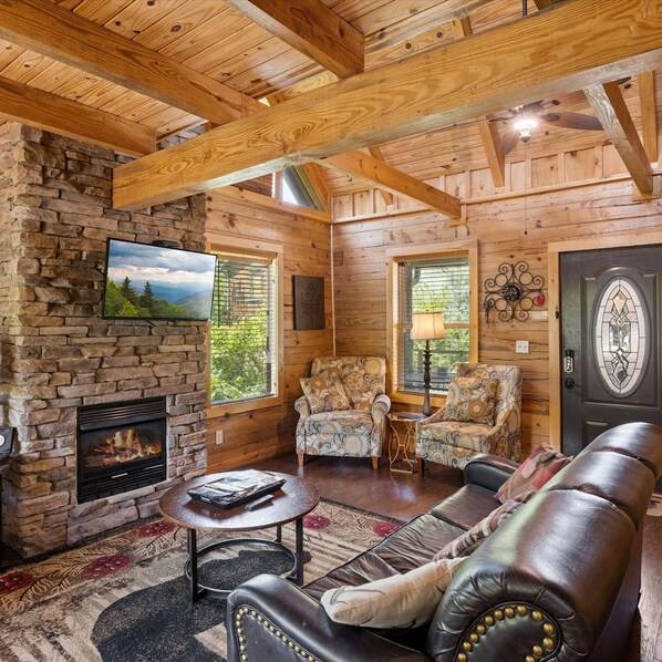 A cozy cabin interior with a stone fireplace, wooden ceiling beams, a leather sofa, and two patterned armchairs. - Pigeon Forge I Hot tub I Theatre I Walk to Parkway (Pigeon Forge)