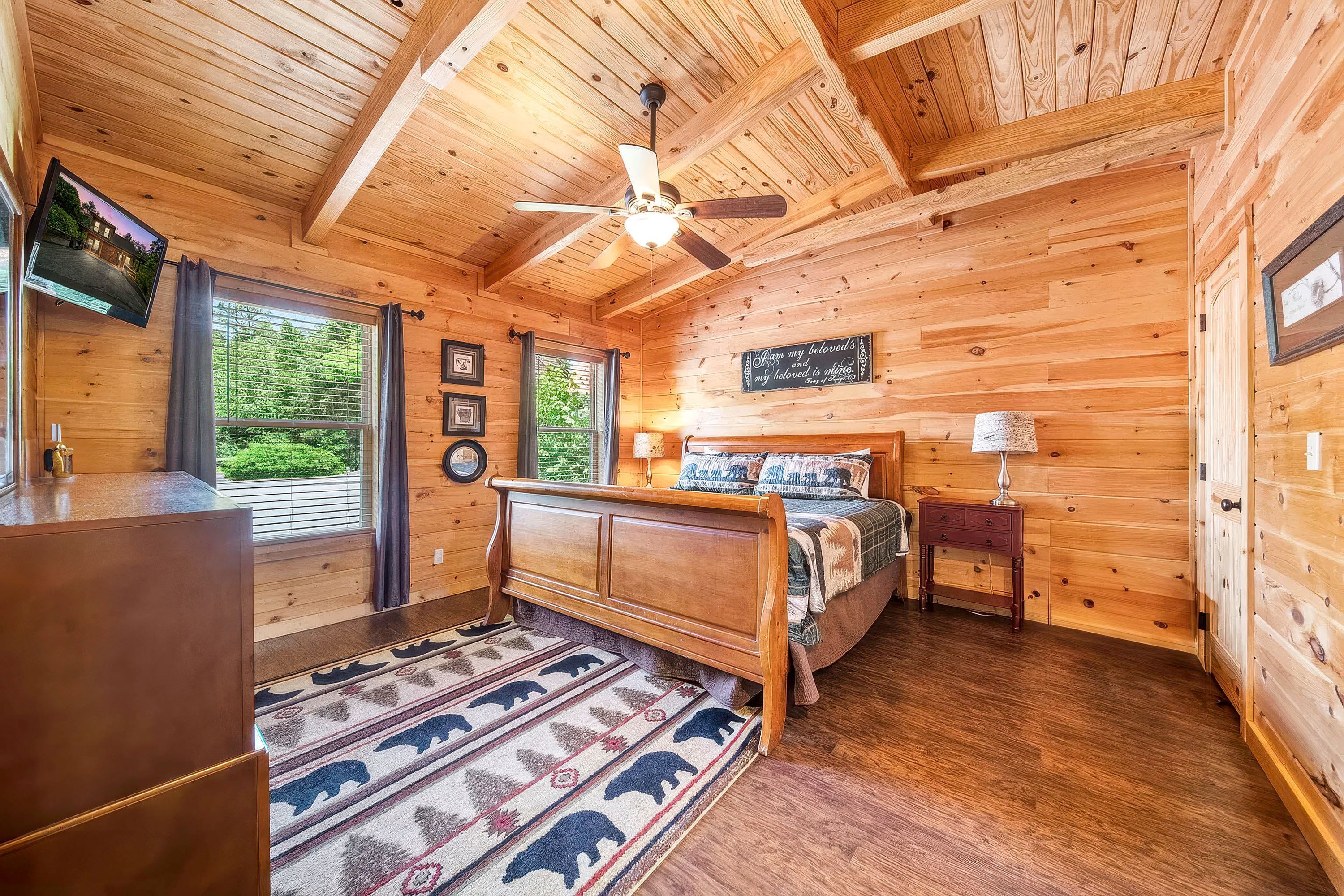 A wooden cabin bedroom with a bed, a ceiling fan, a window with curtains, a wall-mounted TV, and a rug with animal prints.