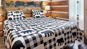 A bedroom with a wooden log wall, a ceiling fan, a bed with a checkered quilt, and a nightstand with a lamp. - Views I Between Pigeon Forge Gatlinburg I Firepit (Sevierville)