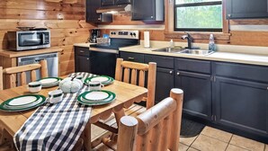 A kitchen with wooden paneling, a dining table with plates and cups, a microwave, a sink, and a window with a view of trees. - Views I Between Pigeon Forge Gatlinburg I Firepit (Sevierville)