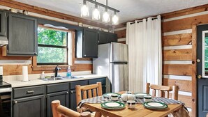 A compact kitchen with dark cabinets, a stainless steel refrigerator, a wooden dining table with chairs, and a window with a view of greenery. - Views I Between Pigeon Forge Gatlinburg I Firepit (Sevierville)