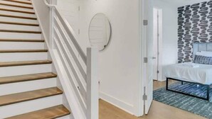 Eine Treppe mit Holzboden und weißen Geländern führt zu einem Schlafzimmer mit Bett, einem gemusterten Wandbild und einem blauen Teppich.