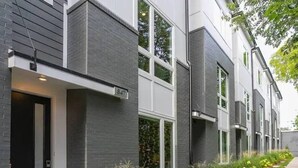 Ein modernes, mehrgeschossiges Wohngebäude mit grauem Backstein-Fassadengestalt, großen Fenstern und einem gepflegten Vorgarten mit verschiedenen Pflanzen.