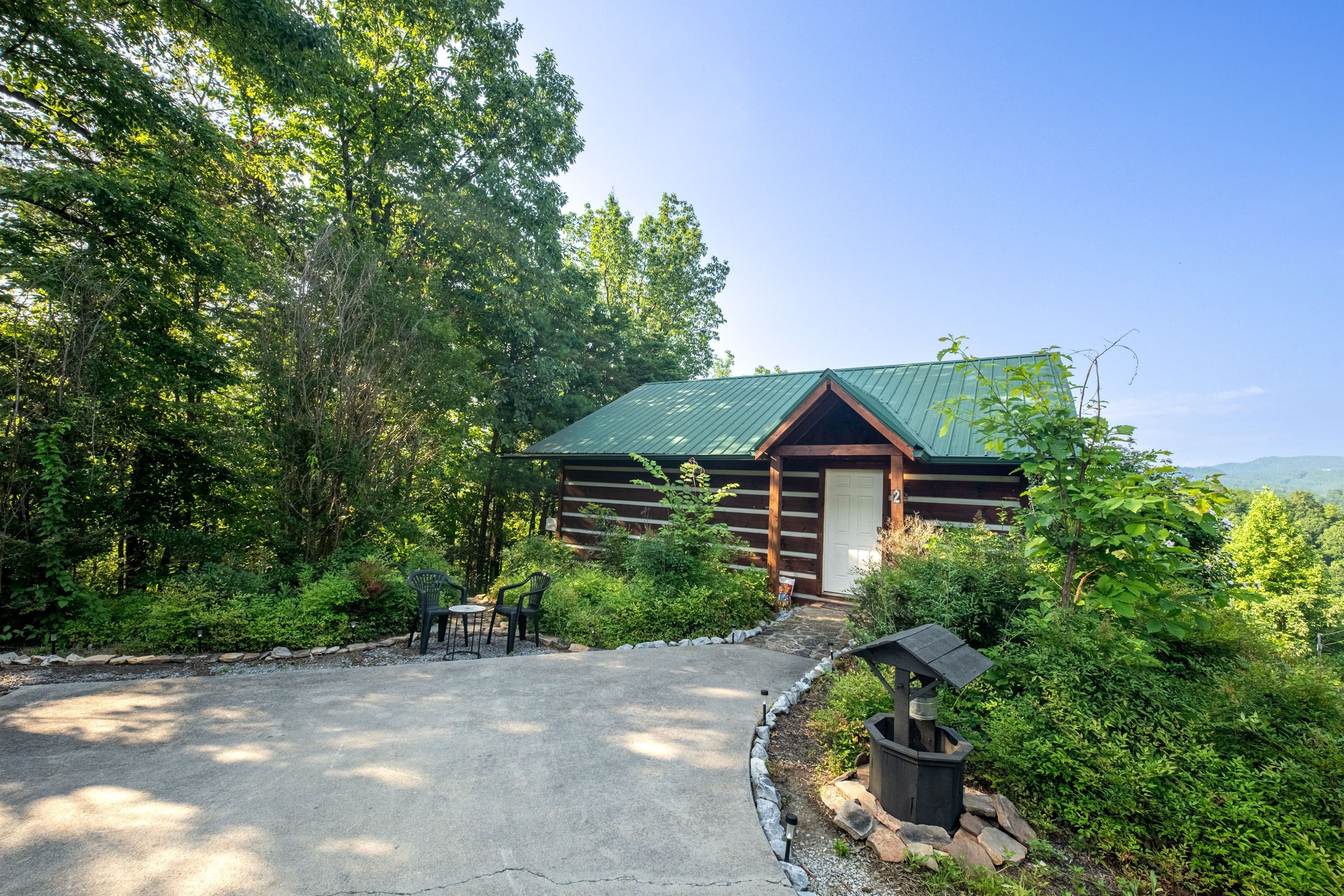 A cabin with a green metal roof, surrounded by dense greenery and a gravel path leading to the entrance.