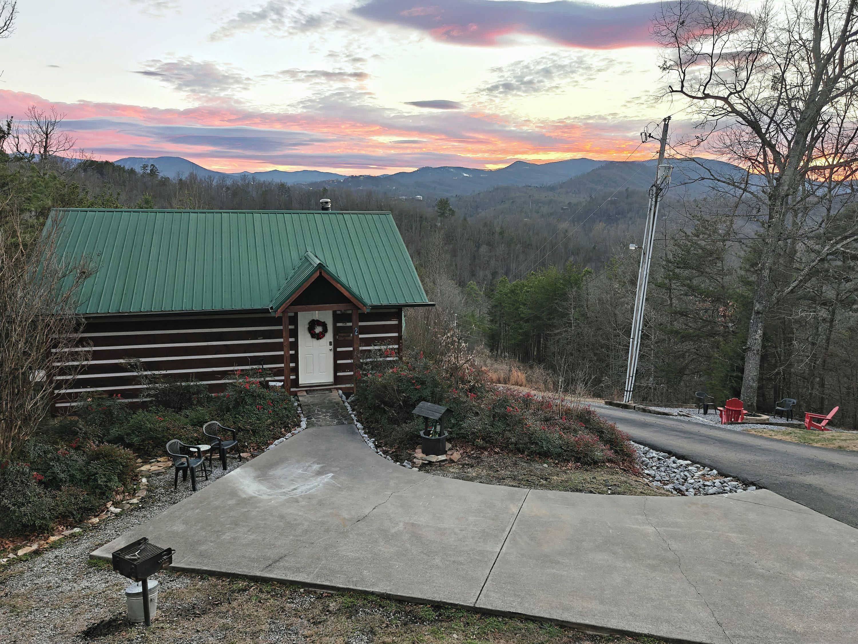 A log cabin with a green roof, a front porch, and a gravel pathway leading to a seating area with outdoor furniture.