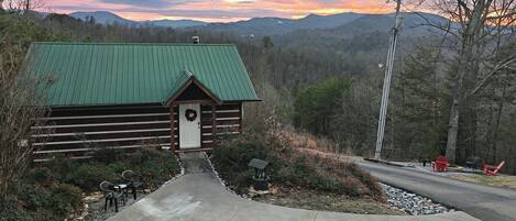 A log cabin with a green roof, a front porch, and a gravel pathway leading to a seating area with outdoor furniture.