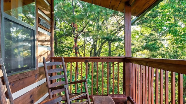 Une véranda en bois, avec un banc et des chaises, donnant sur une zone boisée.