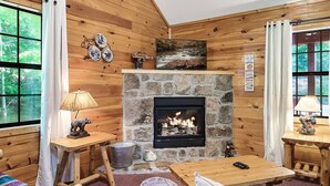 L’intérieur chaleureux d’un chalet avec une cheminée en pierre, des murs en bois et une vue sur la forêt grâce à de grandes fenêtres.