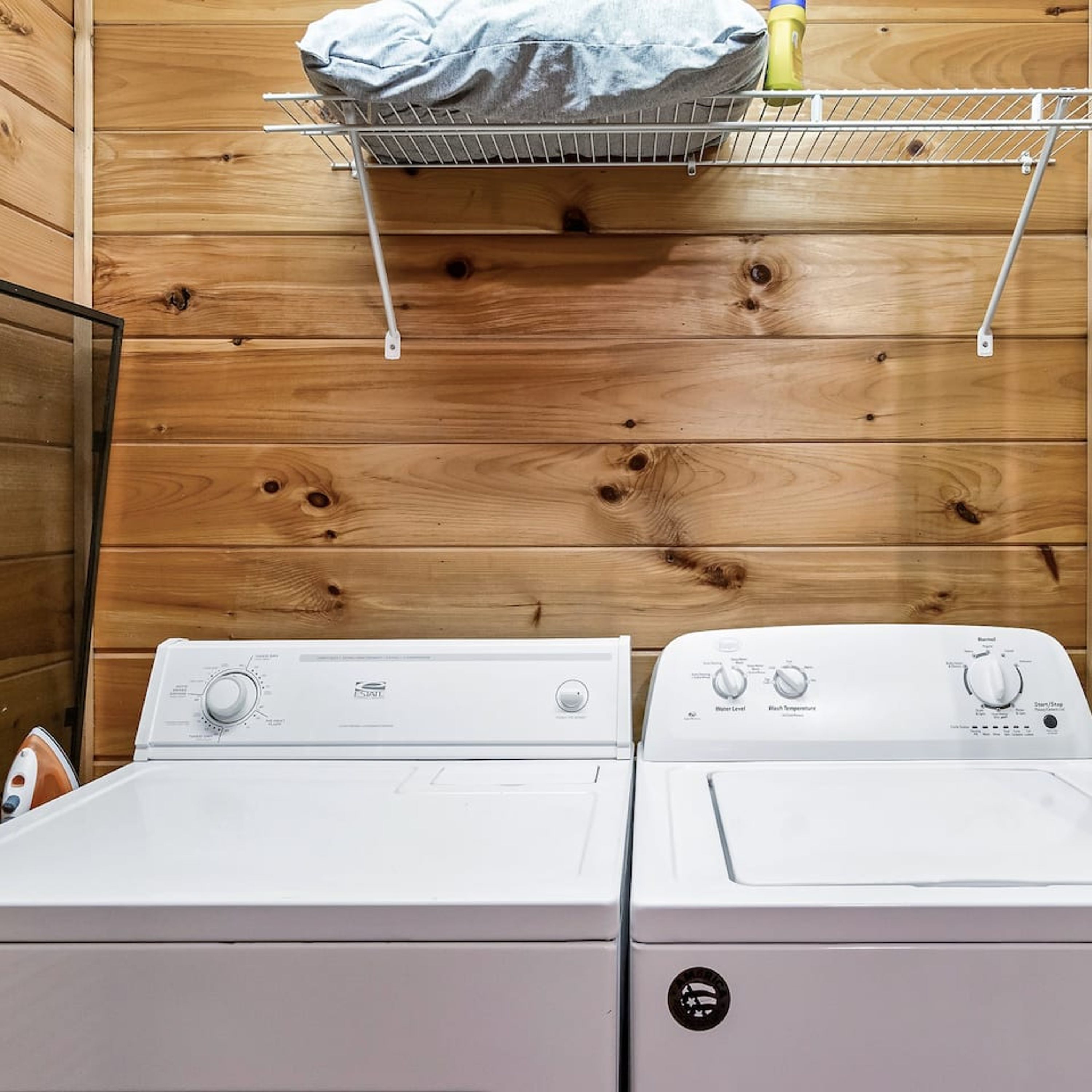 A laundry room with two white washing machines, a wooden wall, and a metal shelf with a blue cloth and a yellow bottle.