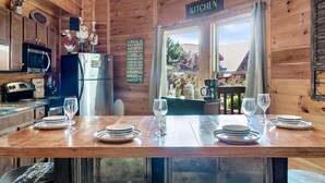 L’intérieur d’un chalet en bois comprend un coin cuisine, une table à manger dressée avec des assiettes et des verres, et une vue sur l’extérieur grâce à de grandes fenêtres.