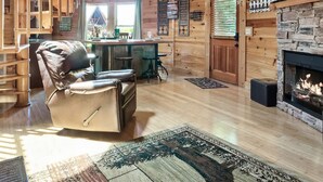 L’intérieur chaleureux d’un chalet, avec un revêtement en bois, une cheminée, un coin cuisine et un espace salon confortable agrémenté d’un fauteuil en cuir.