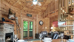 Un intérieur en bois avec une table à manger dressée pour quatre personnes, une cheminée, une horloge et un escalier.