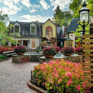 A quaint village square with cobblestone paths, a fountain, and flower beds.