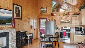 Un intérieur de chalet chaleureux avec une cheminée en pierre, des murs en bois, un coin repas et une cuisine équipée d’appareils électroménagers en acier inoxydable.