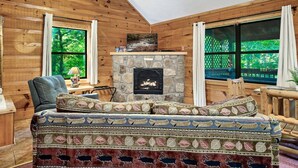 Interior acogedor de una cabaña con chimenea de piedra, paredes de madera, sofá estampado y ventanas amplias con vistas al exterior.