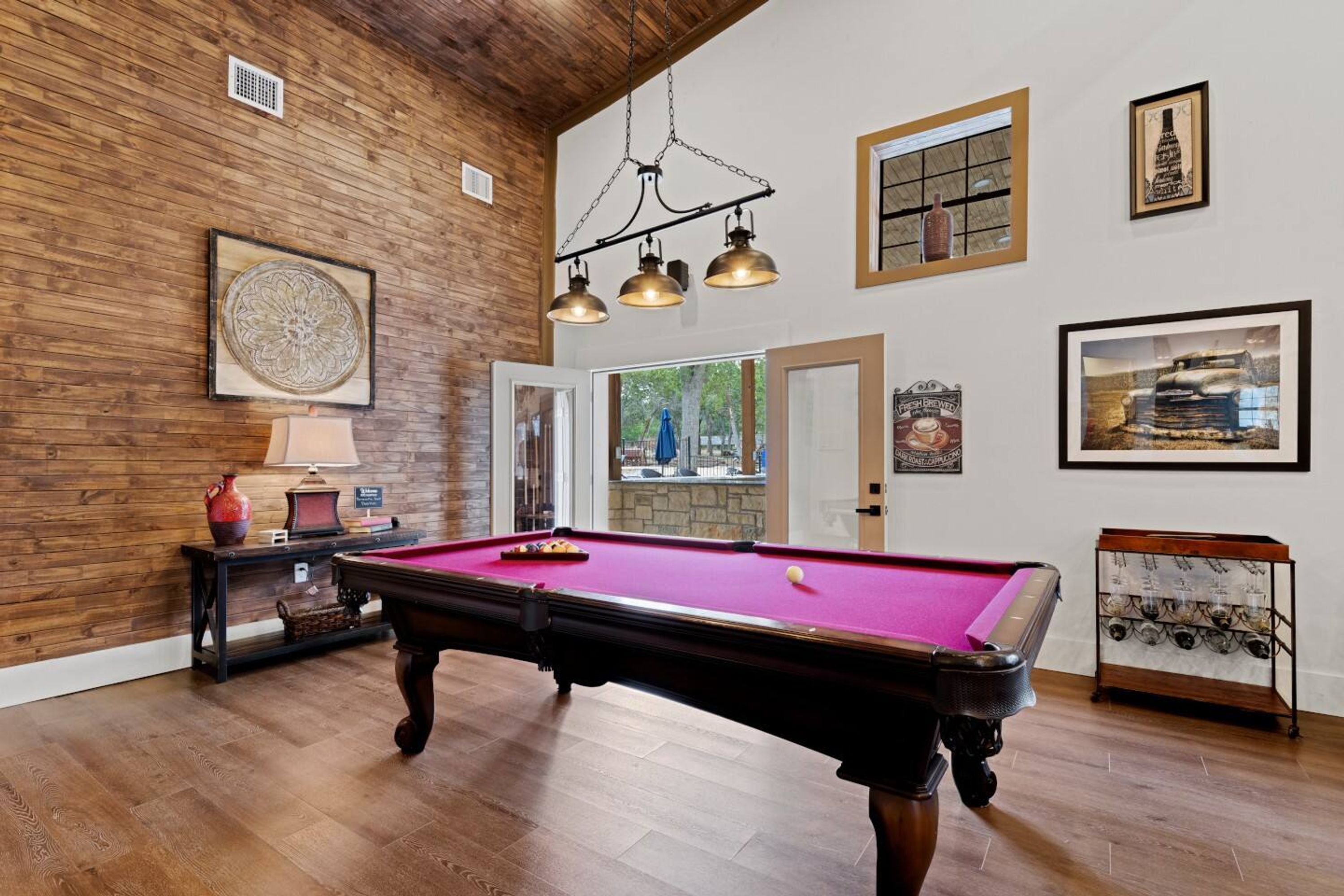 A pool table in a room with wooden walls and a high ceiling. There are framed pictures on the walls and a chandelier above the table.