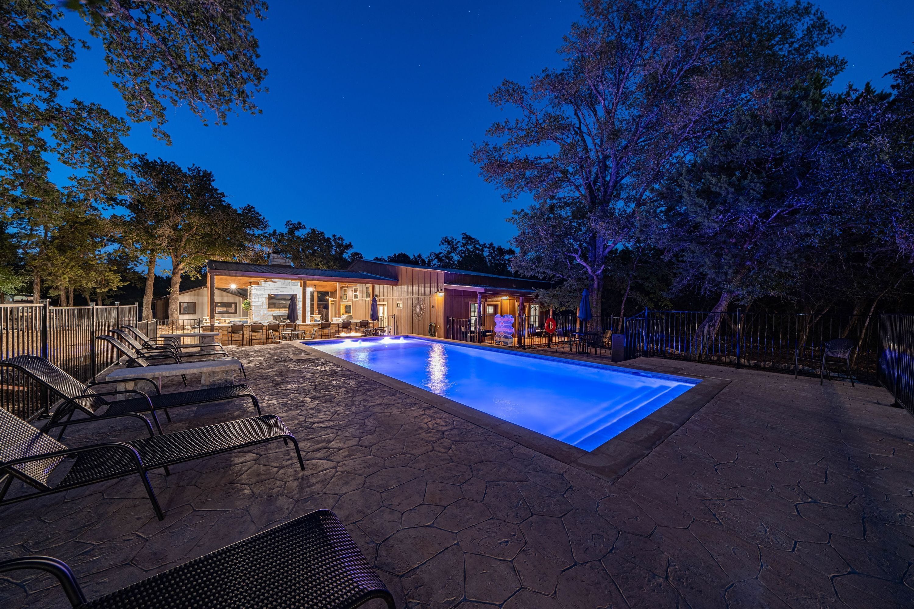 A swimming pool with lounge chairs, a covered patio area, and a building with illuminated windows.