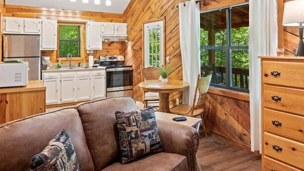 Acogedor interior de cabaña con detalles de madera, una kitchenette, un sofá de cuero marrón con cojines estampados y un comedor con mesa y sillas.