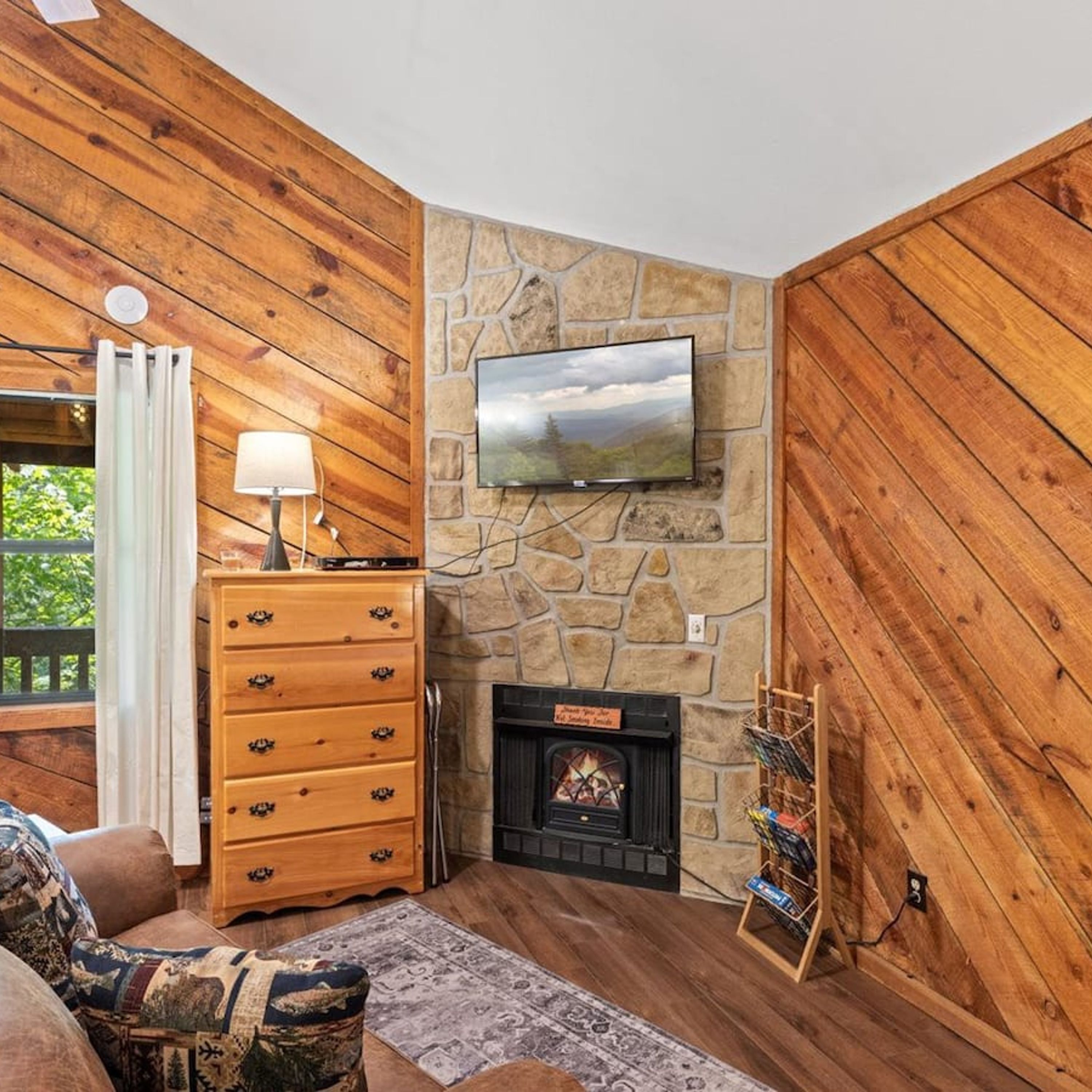A cozy room with wooden walls, a stone fireplace, a television mounted on the wall, and a wooden dresser with drawers.