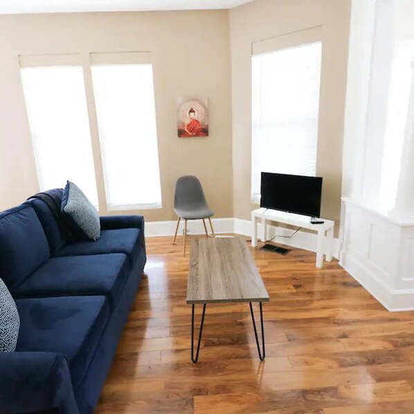 Sala de estar com um sofá azul, uma poltrona cinza, uma mesa de centro de madeira e uma televisão sobre um suporte branco.