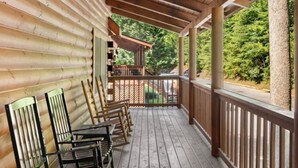 A wooden cabin with a covered porch, wooden chairs, and a view of the surrounding forest.