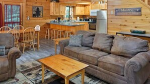 A cozy cabin interior with a wooden staircase, a dining area with chairs, a sofa, and a kitchen with a bar.