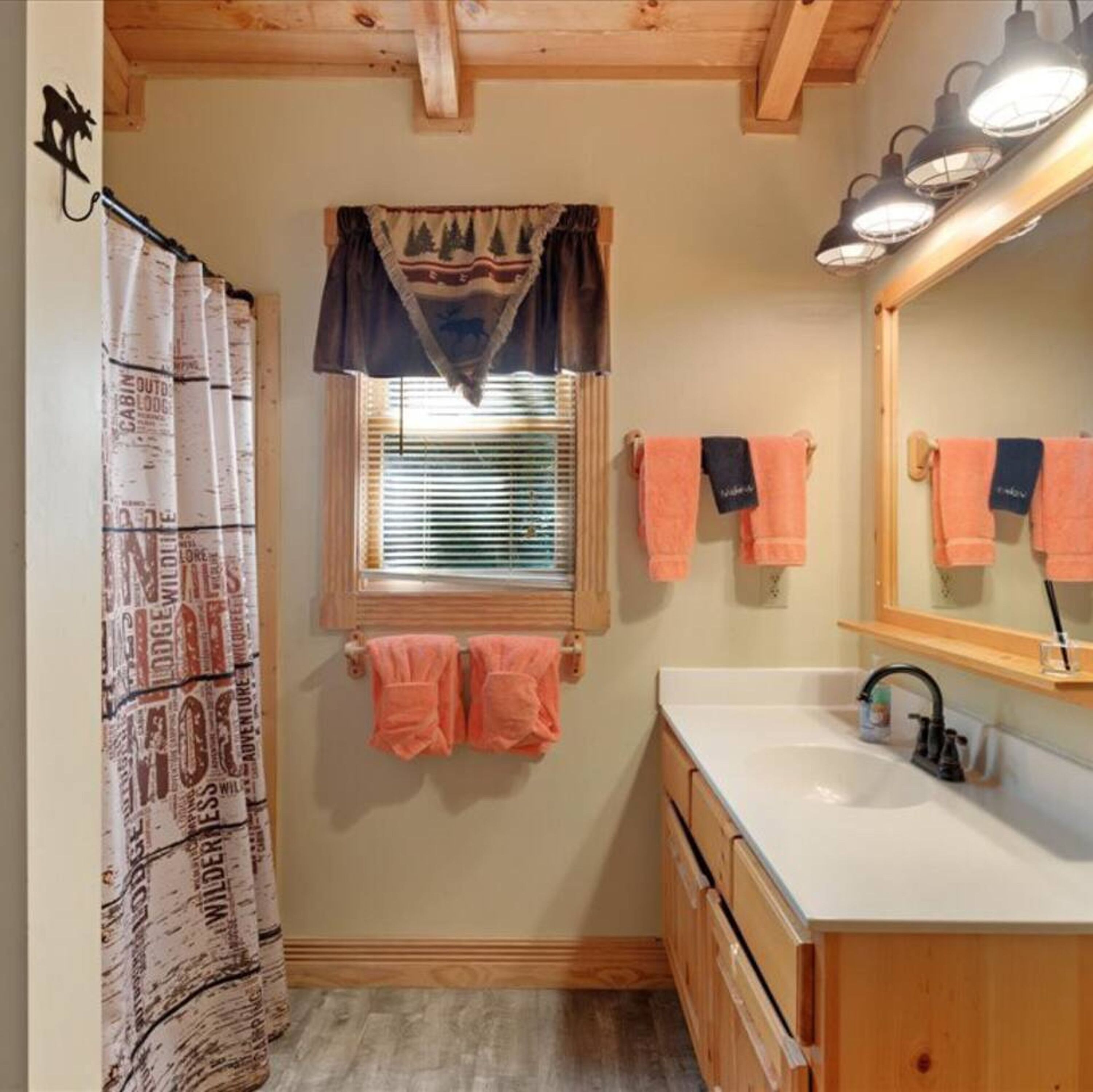 A bathroom with a wooden ceiling, a window with blinds, a sink with a mirror, and towels hanging on the wall.