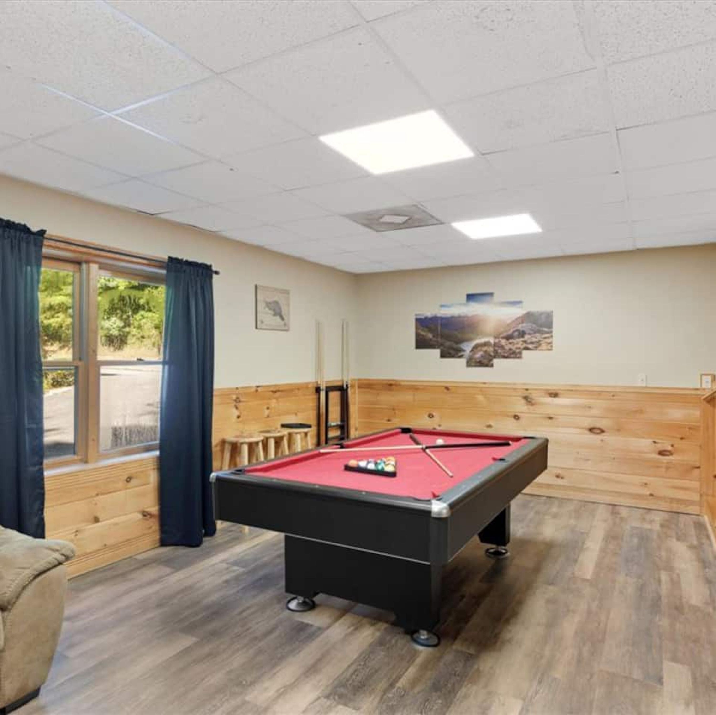A pool table in a room with wooden paneling, a window with curtains, and a wall-mounted picture.
