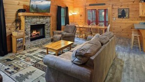 A cozy log cabin interior with a stone fireplace, a leather hide on the wall, a television, and a dining area.