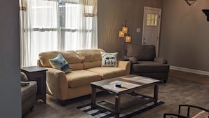 A living room with a beige sofa, a wooden coffee table, and a chair. There is a window with sheer curtains, a lamp, and a door in the background. - 180 Degree Seneca Lake Views, privacy galore with hot tub. Sleeps 9
 (Senecaville)