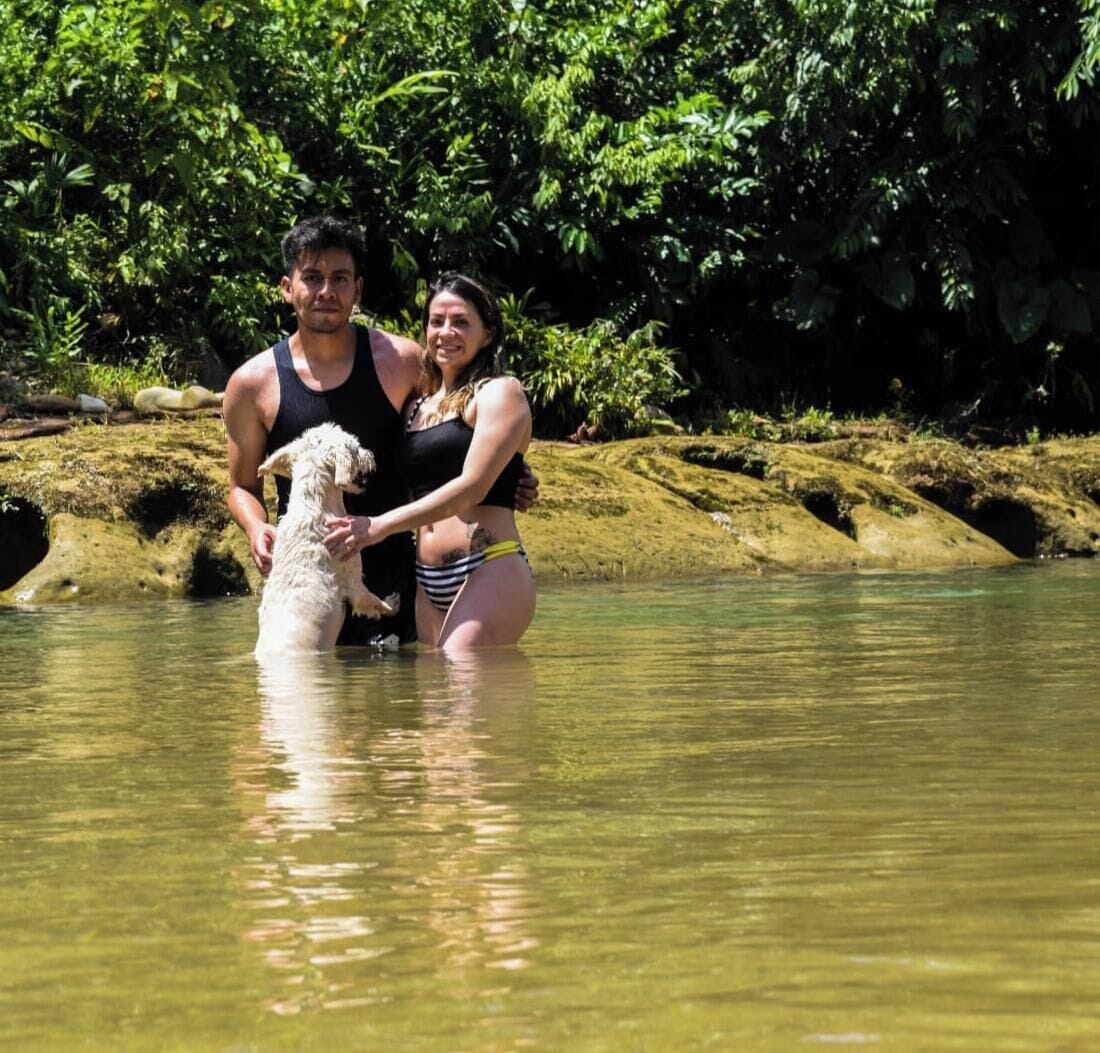 Een stel staat in ondiep water, omringd door weelderige begroeiing, samen met een hond.