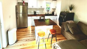 A compact living space with a kitchenette, wooden flooring, a sofa, and two stools.