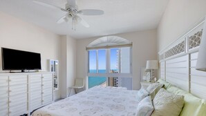Une chambre à coucher comprenant un lit, une télévision, un ventilateur de plafond, une fenêtre donnant sur la mer et une commode.