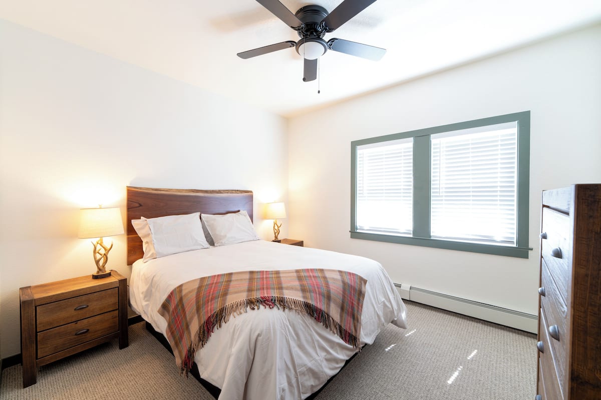 Ein Schlafzimmer mit Bett, Nachttisch, Kommode, Deckenventilator und einem Fenster mit Jalousien.