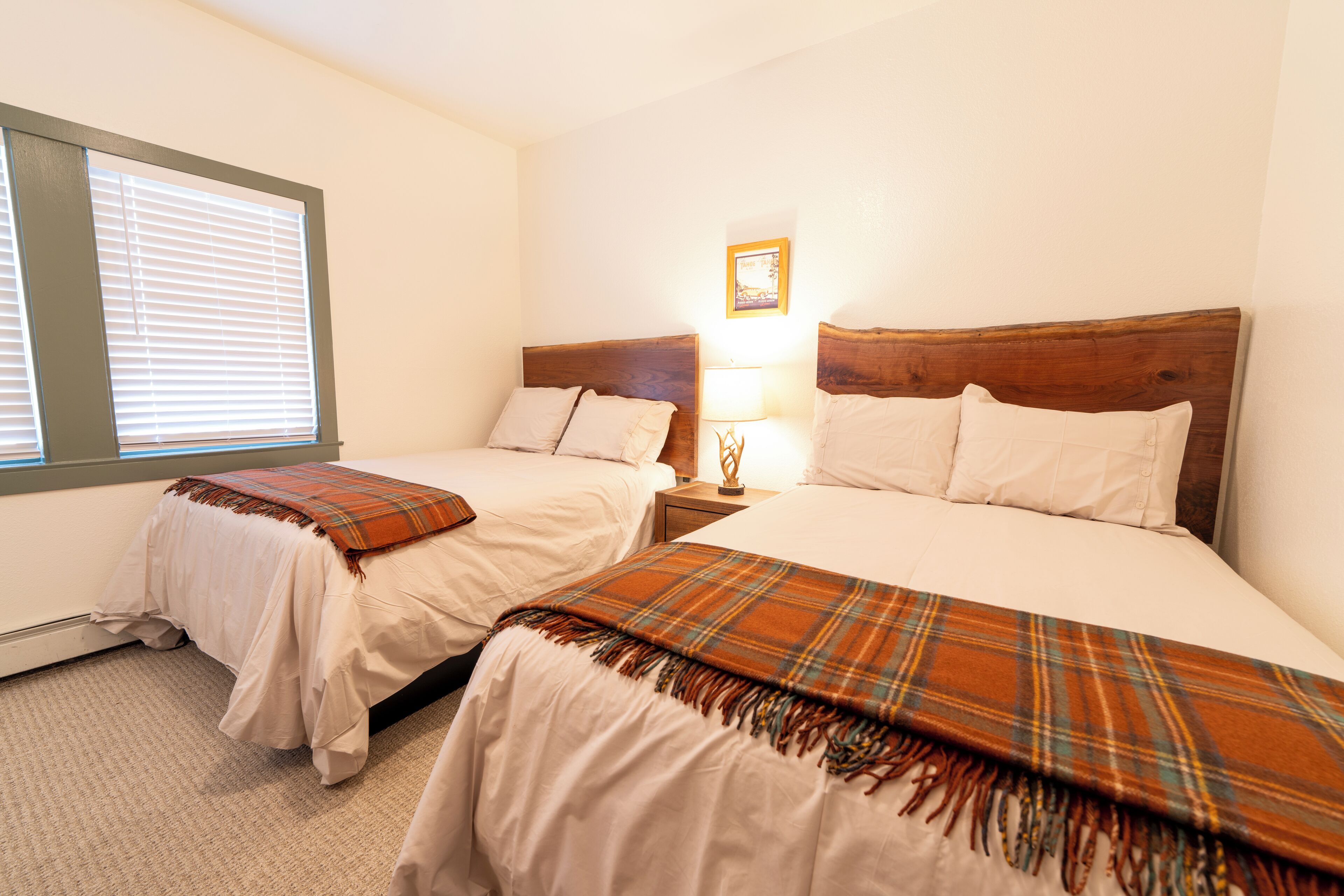 A hotel room with two beds, a window with blinds, a bedside table with a lamp, and a framed picture on the wall.