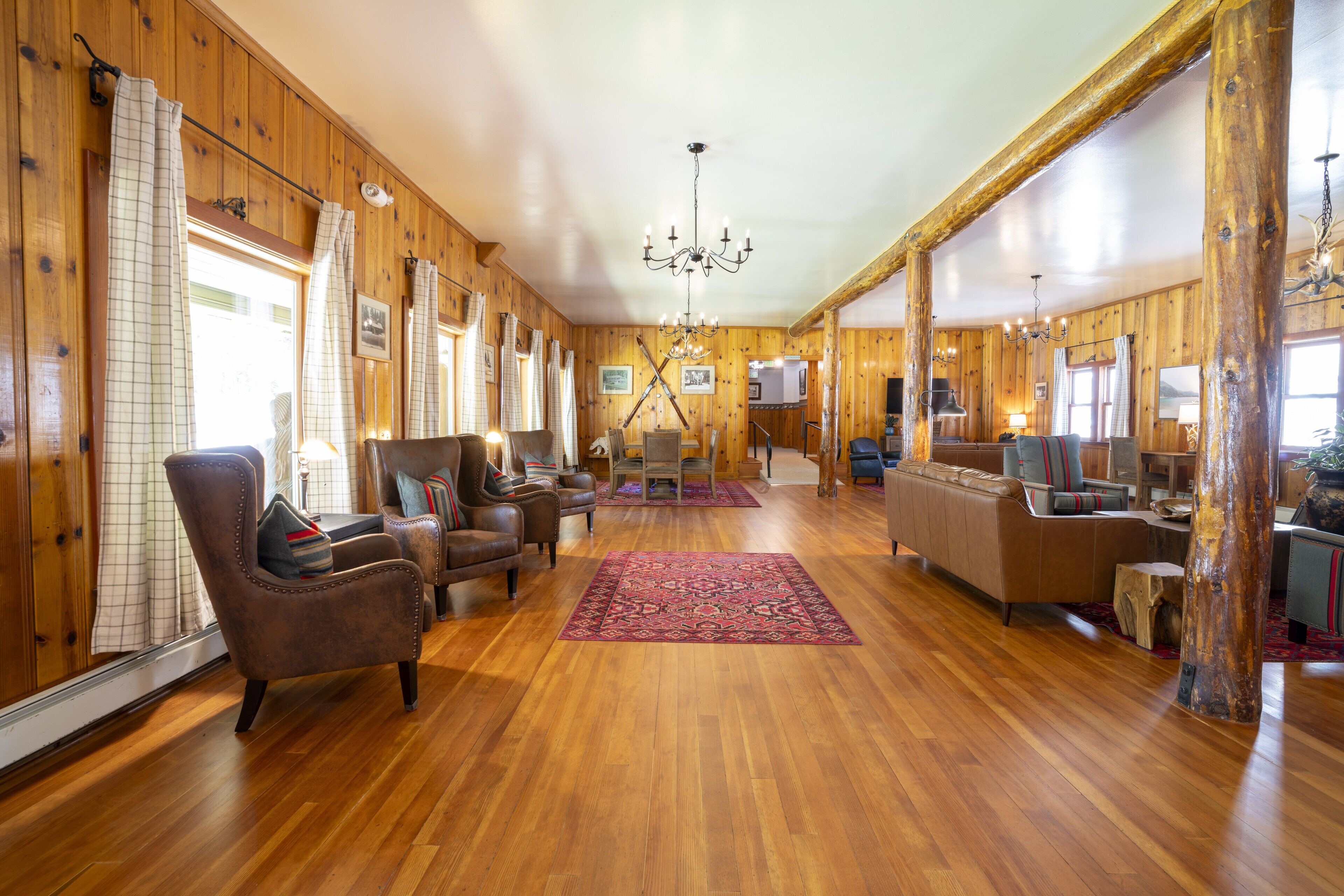 A spacious room with wooden walls and floors, featuring a large chandelier, leather armchairs, a sofa, and a wooden coffee table.