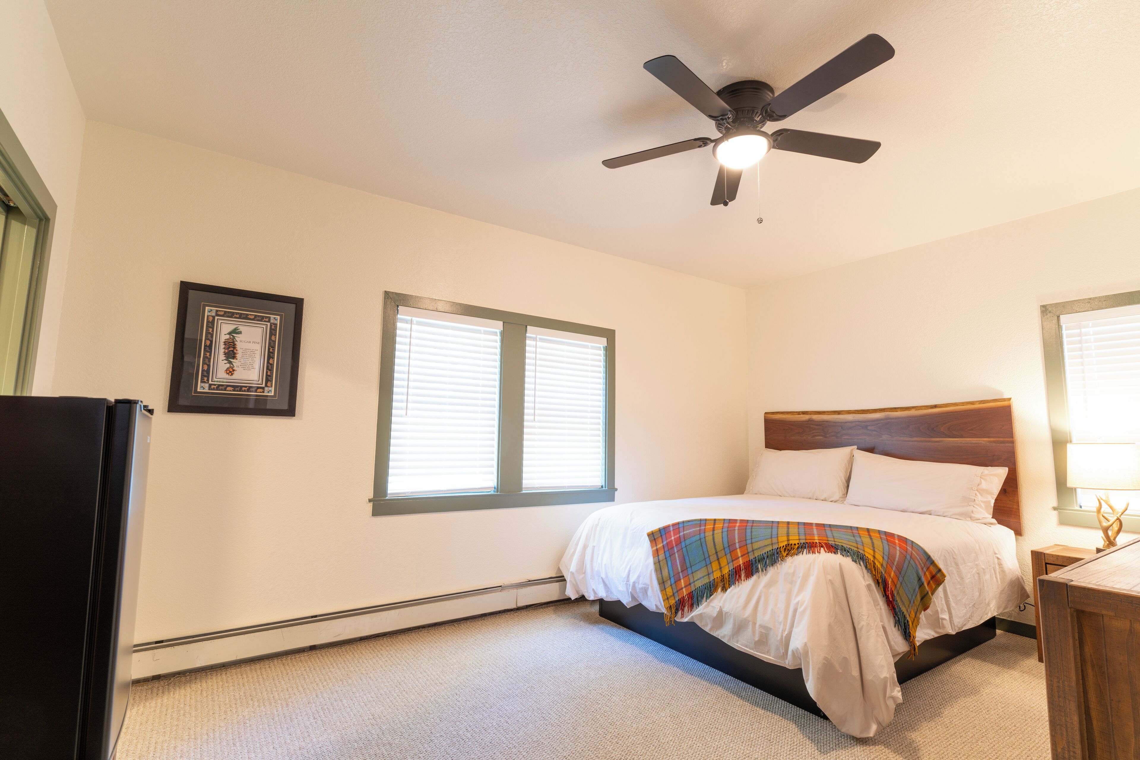 Un dormitorio con cama, ventilador de techo, ventana y un cuadro en la pared.