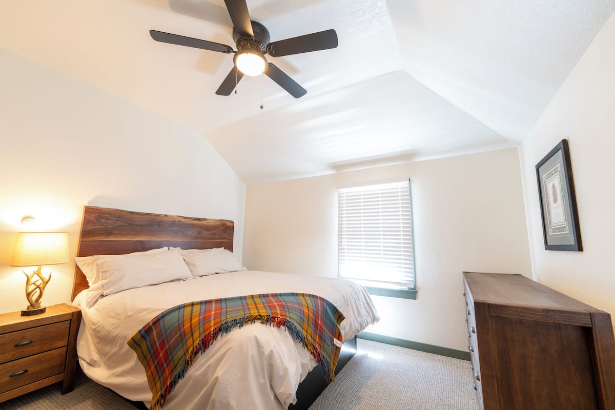 Ein Schlafzimmer mit einem Bett, einem Nachttisch, einer Holzkommode, einem Deckenventilator und einem Fenster mit Jalousien.