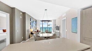A modern hotel room with a large central dining table, a sofa, and a TV. The room has a view of the sea and a balcony.