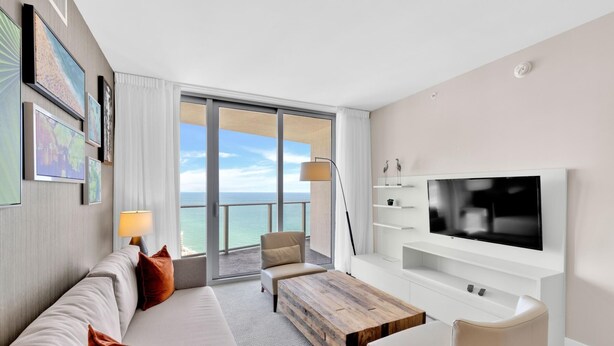 A modern hotel room with a large balcony, a flat-screen TV, a sofa, a coffee table, and a TV stand with shelves.