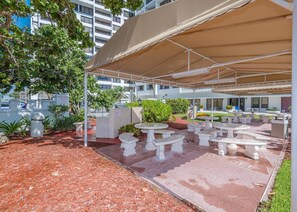 Une terrasse extérieure avec des tables et des chaises en pierre blanche, un espace couvert doté d’un auvent beige, et une vue sur un bâtiment moderne en arrière-plan.
