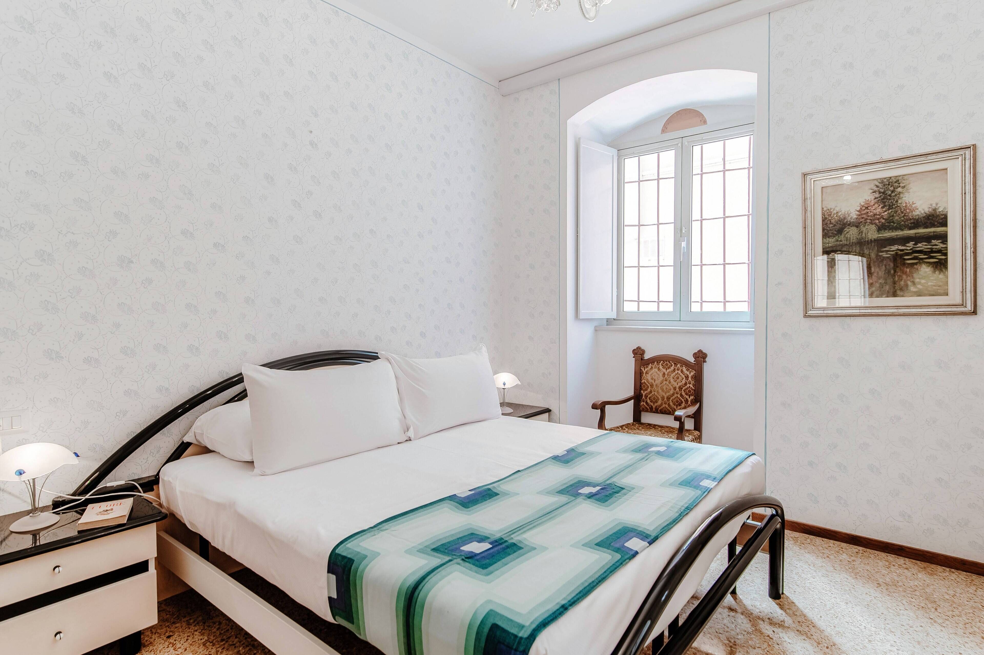 A neatly made bed with a patterned bedspread, a bedside table with a lamp, and a chair against the wall.