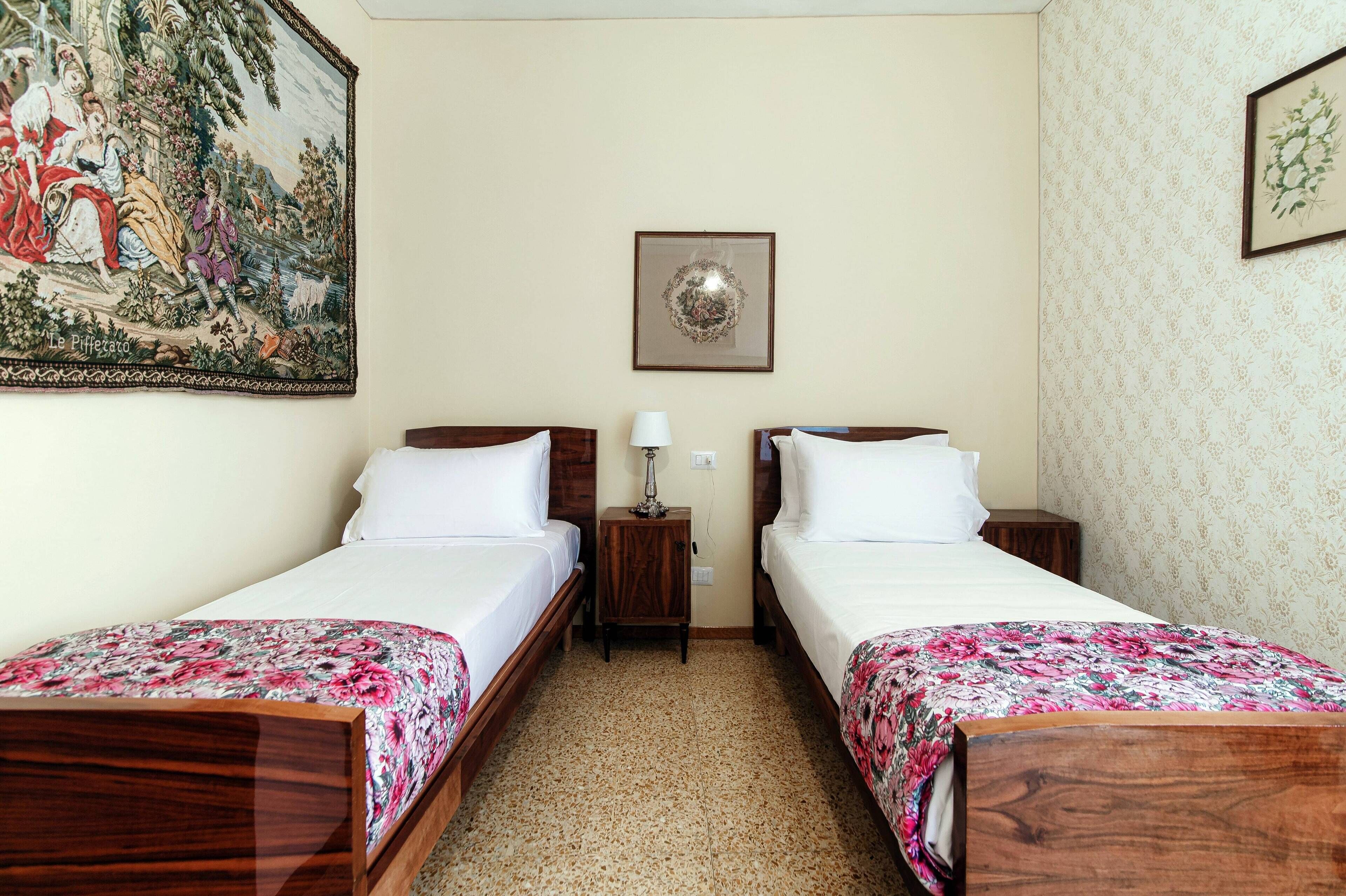 Una camera d'albergo con due letti singoli, un comodino in legno con una lampada, un quadro incorniciato sopra i letti e una moquette con motivi floreali.
