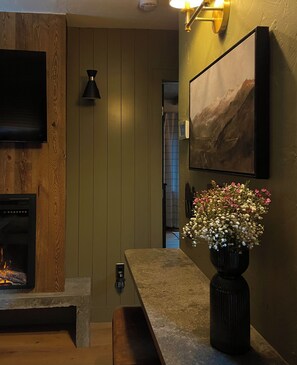 Una sala de estar acogedora con chimenea, una pared de madera, un banco de piedra, un jarrón con flores, un cuadro y una lámpara de pared.